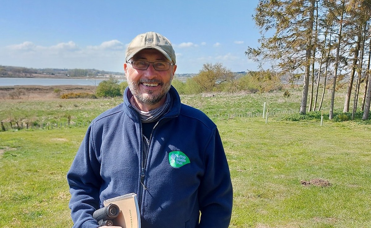 Graham Hart smiling outdoors, wearing a Suffolk Wildlife Trust jacket and cap, with trees and fields in the background